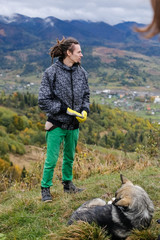 Young man with dreadlocks stands before a dog lying on the hill