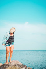 Tanned blonde woman walking at summer beach. Mature female in denim shirt, standing on rocky coast, blue sky background with sea. Concept holiday, outdoors. Active lifestyle.