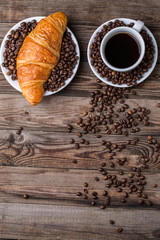 Breakfast coffee - cup and croissant with coffee beans on wooden background.