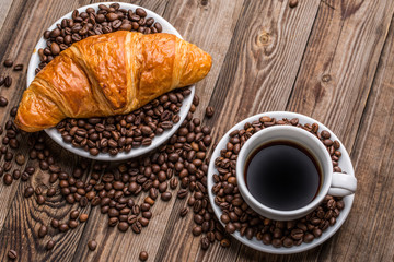 Breakfast coffee - cup and croissant with coffee beans on wooden background.