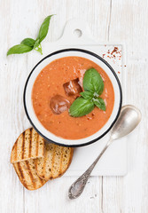 Cold gazpacho tomato soup in bowl with ice, hot pepper and basil served with toasted bread on ceramic board over white painted wooden background, top view, vertical composition