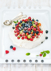 Homemade Pavlova cake with fresh garden berries served with silver spoons on white baking tray over white painted wooden background, top view, selective focus, vertical composition