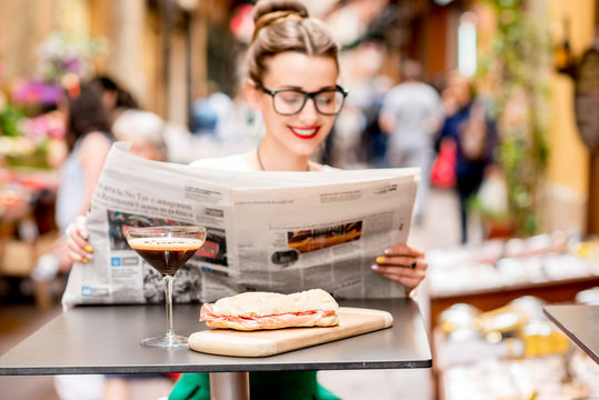 Young woman reading newspaper with shakerato drink and panini on the table outdoors on the street in italian city. Traditional italian lunch. Image with small depth of field, focused on the food