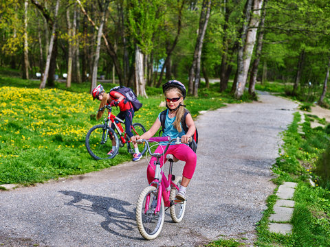 Bikes bicyclist girl. Two sister girls wearing bicycle helmet and glasses with rucksack rides bicycle. Bicyclist children is looking camera. Children sisters ride on green grass and flowers in park.