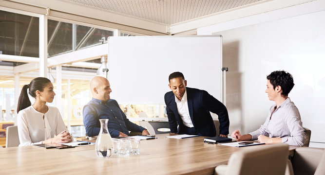 Businessman taking a seat at the head of a board room table next to one of his female colleagues, ready to collaberate with all 3 other business executives that were already seated, waiting and ready