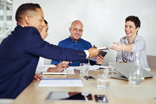 Group of four business executives in the middle of a board meeting in the conference room, everyone looking happy as a mature business woman passes a notebook to her business partner.