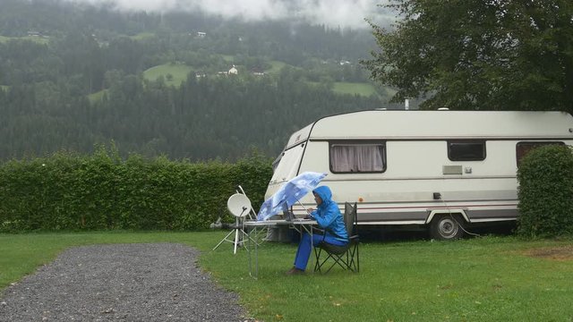 A woman works on laptop under blue umbrella under rain in camping near trailer