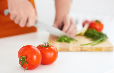 Two ripe tomatoes in the foreground . Man is cutting ripe vegetables on a wooden board in the background.