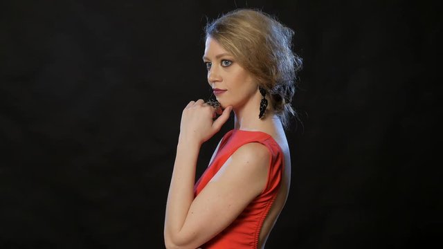 pretty young blonde standing half-turned on black background, touching hair and earring