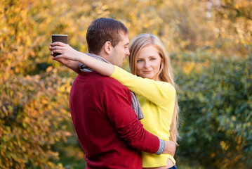 Fototapeta premium Love, relationship, season, friendship and people concept - happy man and woman enjoying golden autumn fall season with paper coffee cups in the park. Young couple drinking hot cofee or tea.