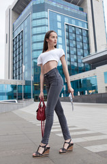 Naklejka premium Beautiful brunette young woman wearing black jeans, white shirt and walking on the street . Fashion magazine in hand. Blurred background.