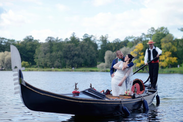 Wedding couple is hugging on a floating gondola. Beauty bride with groom. Beautiful model girl in white dress. Man in suit. Female and male portrait. Woman with lace veil. Cute lady and guy outdoors