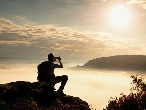 Dreamy fogy landscape. Amateur photographer takes impressive photos with phone. Hiker with backpack on cliff photogrphy  orange pink misty sunrise in a beautiful valley below