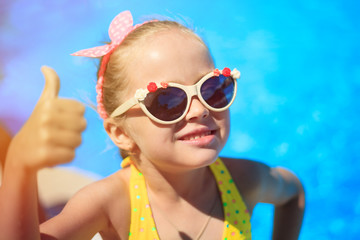 Little girl showing thumbs up on the pool background. Happy smiling Child is wearing sunglasses, enjoying pool and having fun. Travel (vacation), adventure concept. Close up, outdoor.