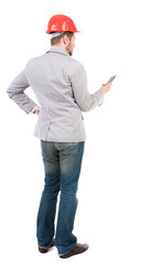 Backview of business man in construction helmet stands and enjoys tablet or using a mobile phone. Standing young guy. Rear view people collection.  backside view of person.  Isolated over white