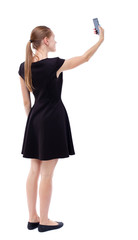 back view of standing young beautiful  woman  using a mobile phone. girl  watching. Rear view people collection.  backside view of person.  Isolated over white background. Blonde in a short black