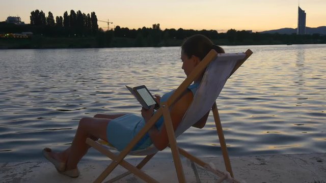 Female sits on a sunbed reading e-reader at the waterfront