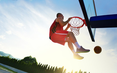 Young basketball player drives to the hoop for a high flying slam dunk in front of sunset sky.