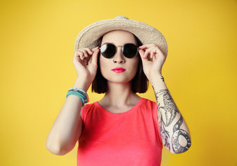 Beautiful young woman with tattoo and sunglasses wearing hat on yellow background
