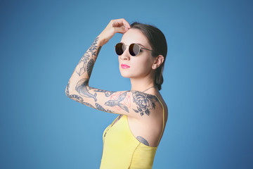 Beautiful young woman with tattoo wearing sunglasses and posing on blue background