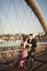 Fototapeta premium Stylish loving wedding couple, groom, bride with pink dress kissing and hugging on a bridge at sunset