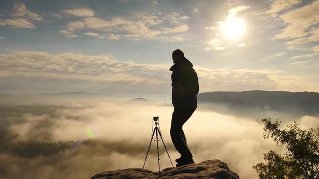 Photographer in green windcheater and black trekking trousers stay at camera on tripod close to cliff edge. Dreamy fogy landscape, blue misty sunrise in a beautiful valley below
