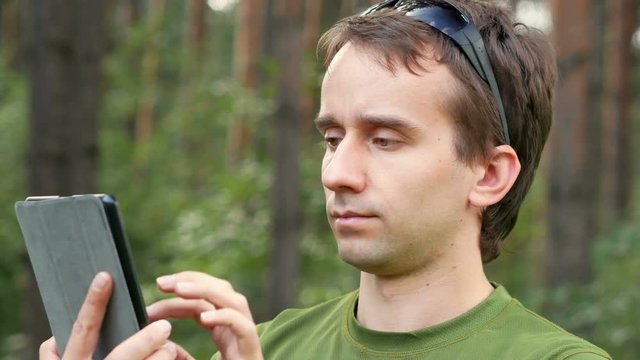 Handsome middle-age man in gren t-shirt sitting touch a tablet. He is smiling,summer forest on the background. Close-up