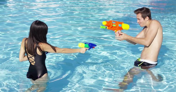 Happy young male and female couple shooting off colorful plastic water guns at each other in swimming pool