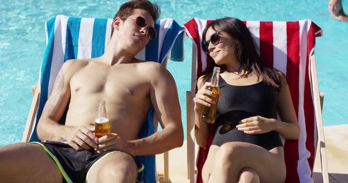 Attractive young couple enjoying a beer while relaxing in their swimsuits on deck chairs near a pool toasting each other with the bottles
