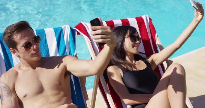 Man and woman taking self portraits with their smart phones at while seated in deck chairs at the swimming pool outside