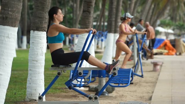 Young athlete woman is doing some sport exercises on rowing machine in the park near the beach