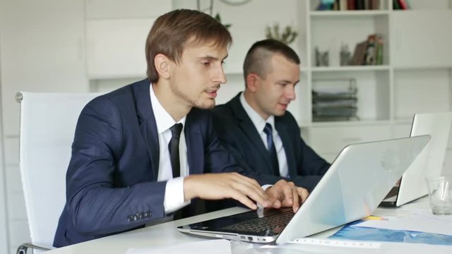  

two glad business male assistants wearing formalwear working together using laptops in company office