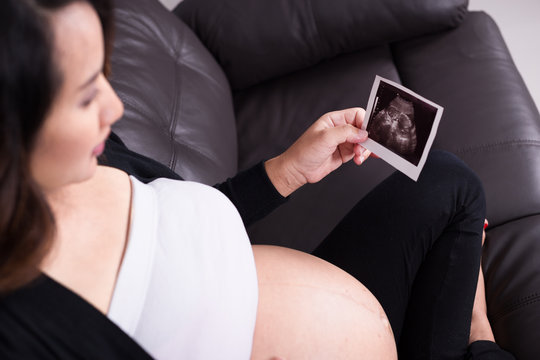 Pregnant woman sitting on sofa, holding her child ultrasound pic