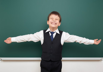 school student boy posing at the clean blackboard and open arms, grimacing and emotions, dressed in a black suit, education concept, studio photo