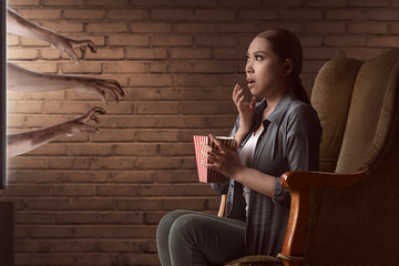 Asian young woman watching horror movie and eat the popcorn with sitting on sofa