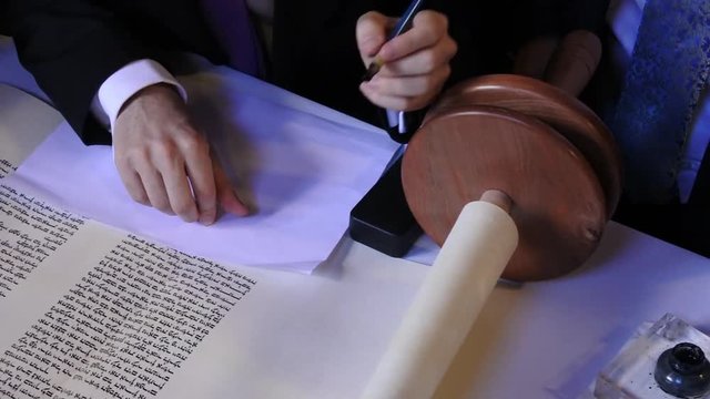 Person holds sofer hands while he completing the final letters of sefer Torah.One can fulfill the last of the 613 commandments mitzvah by commissioning the writing of a scroll.