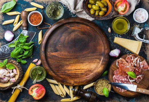 Italian food cooking ingredients on dark plywood background with round wooden tray in center, top view, copy space, horizontal composition