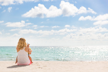 Back view on mother with a small adorable baby sitting on the sandy ocean beach. Family on...