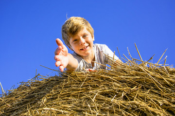 Naklejka na ściany i meble Smiling boy on a haystack on the background of blue sky pulls the arm to the camera