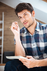 Pensive young man writing in notepad and thinking at home