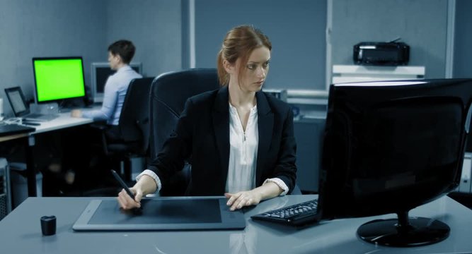 4K: Eine Geschäftsfrau arbeitet an einem Grafiktablet in einem Büro. Eine andere Geschäftsfrau arbeitet im Hintergrund am Pc mit einem Greenkey Monitor. Szene Blaeulich.