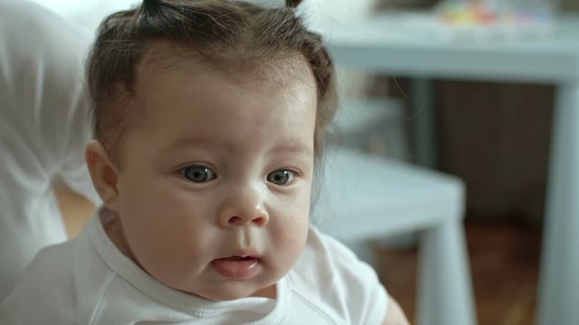 Lovely little baby girl with chubby cheeks sitting with mom and looking at something with open mouth