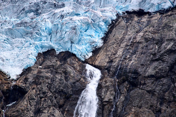 Blue ice and a small waterfall with melted water at the edge of Folgefonna glacier on a mountain...