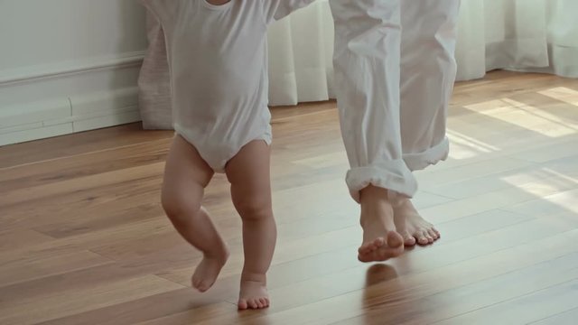 Tilt up of mother holding hands of little baby girl and helping her to walk barefoot in the living room