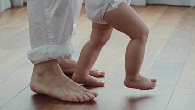 Side view of barefoot legs of woman walking on the floor behind little baby in diaper 