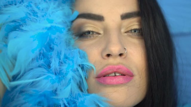 Closeup portrait of beautiful seductive brunette woman smiling and looking into the camera while playing with blue feather boa over blue wall background
