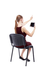 back view of pointing woman sitting on chair and looks at the screen of the tablet.  Rear view people collection.  backside view of person.  Isolated over white background. A girl in a burgundy dress