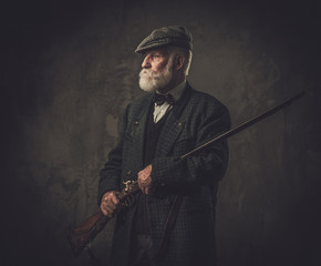 Senior hunter with a shotgun in a traditional shooting clothing, posing on a dark background.