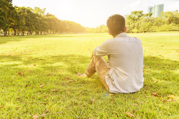 short hair man is relaxing under tree shadow on grass field with nature and sunlight at public park. copy space for message concept of relax. Warm tone sunlight effect.