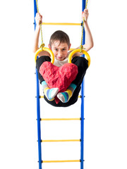 Little boy hanging on sports rings and keeps kicking a red heart on a white background